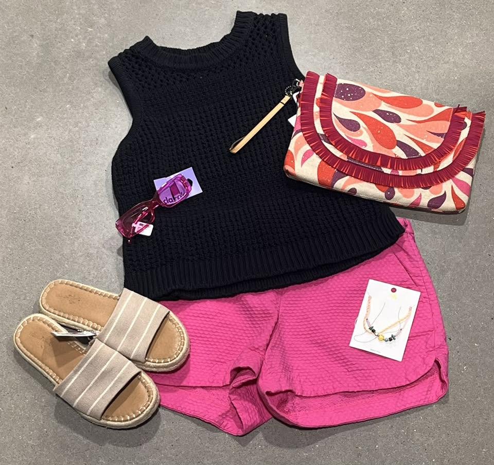 Flat lay outfit with a black knit top, pink shorts, woven sandals, a printed clutch and sunglasses